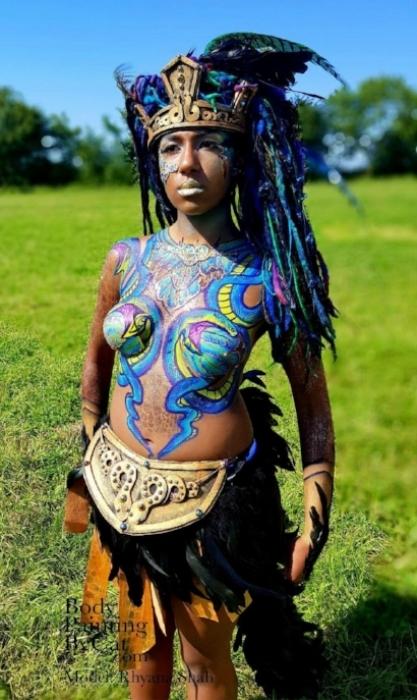 Rhyana in a grass field with warrior outfit, feathered headdress and snake-theme chest body art.