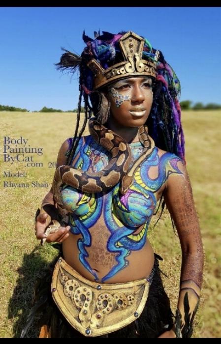 Rhyana in golden warrior's outfit with snake-themed body art, holding a brown and black live snake.