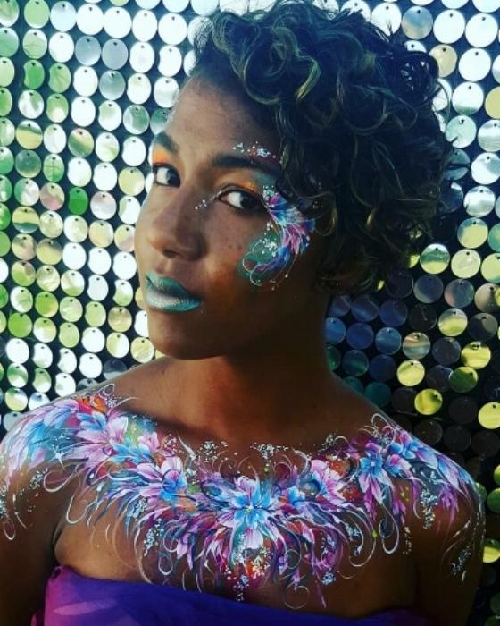 Rhyana with floral rainbow body art on their chest and face, against a mirrored background.
