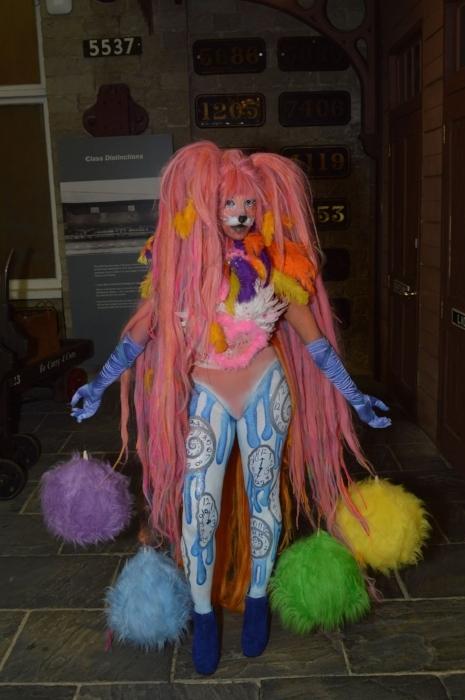 Rhyana in a colourful fox costume with a wig of long pink hair, with colourful fur tassles dangling from their arms.