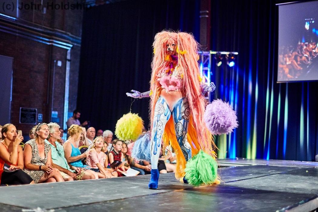 Rhyana in a colourful fox costume, walking on a modelling catwalk with colourful fur tassles dangling from their arms.