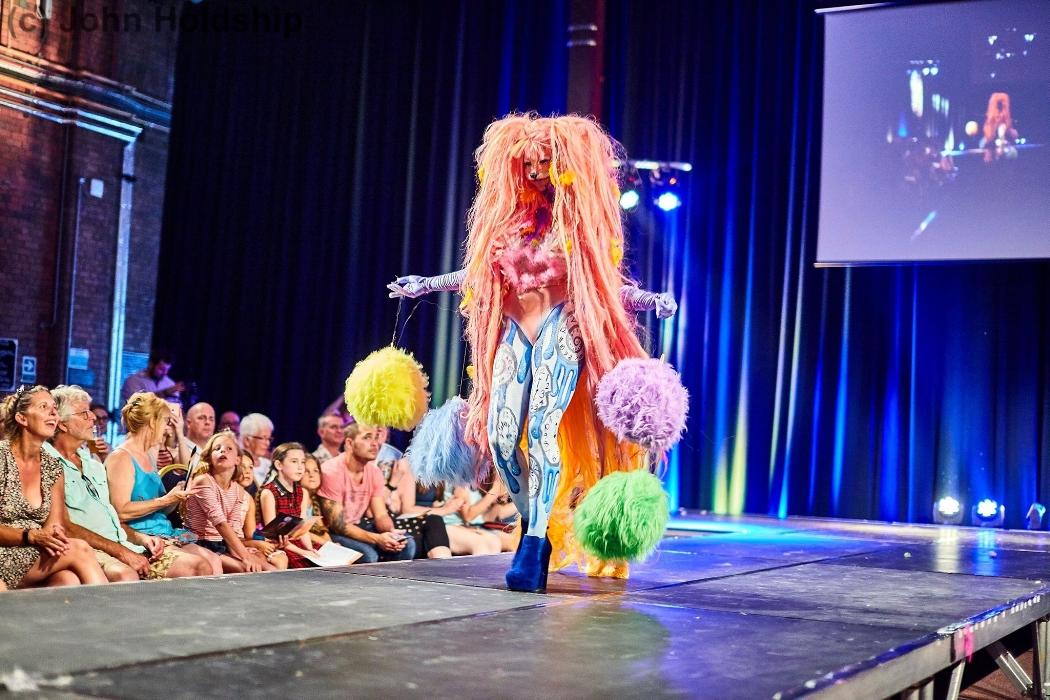 Rhyana in a colourful fur fox outfit with blue heels, walking on a modlling catwalk.