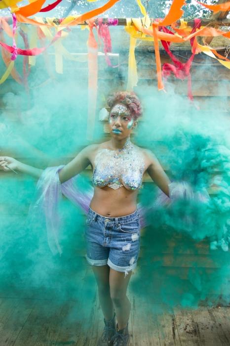 Rhyana posing in denim shorts and silver body paint, surrounding by green smoke.