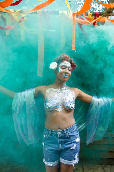 Rhyana smiling in denim shorts, silver body paint and a turquoise shawl, surrounded by green smoke.