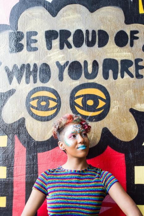 Rhyana in a multi-colored striped t-shirt, smiling against a wall which reads 'be proud of who you are'.