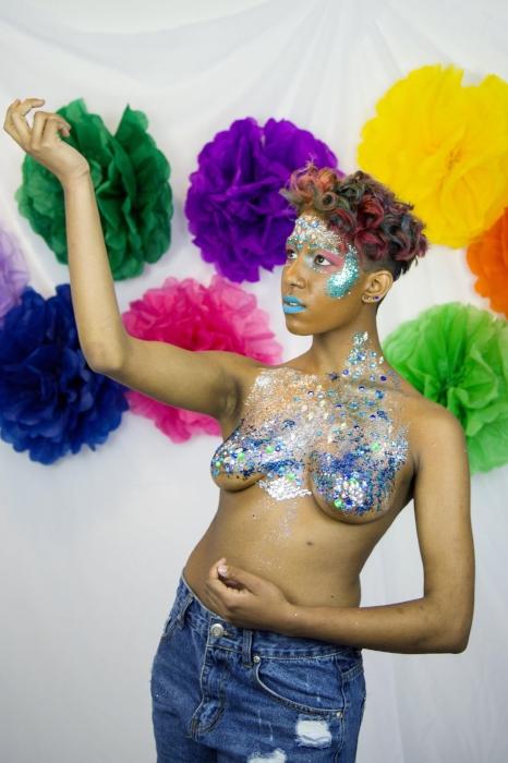 Rhyana posing topless, with arms raised in denim shorts and silver body paint.