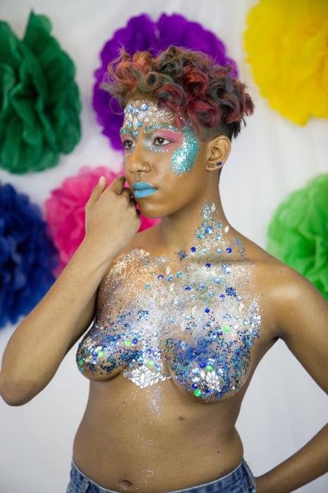 Rhyana looking off camera with one hand on their cheek, dressed in denim shorts and silver / blue bodypaint.