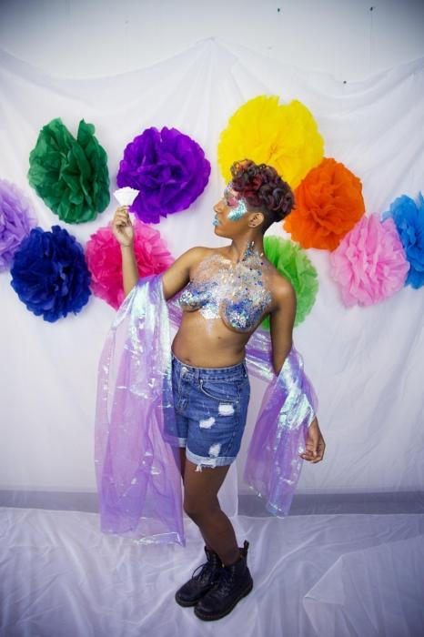 Rhyana posing in denim shorts, silver bodypaint and a purple shawl, stood in front of a wall of multicolored fabrics.