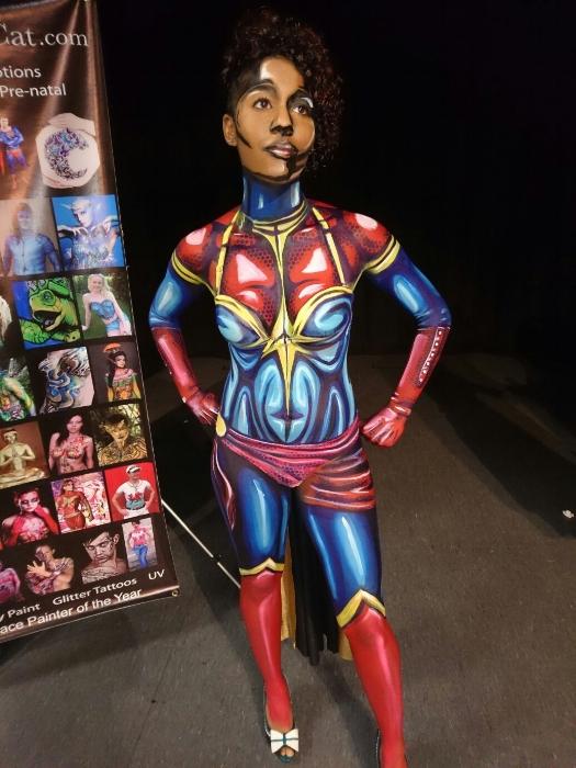 Rhyana posing with body painted as Ms. Marvel, next to a body paint convention poster.