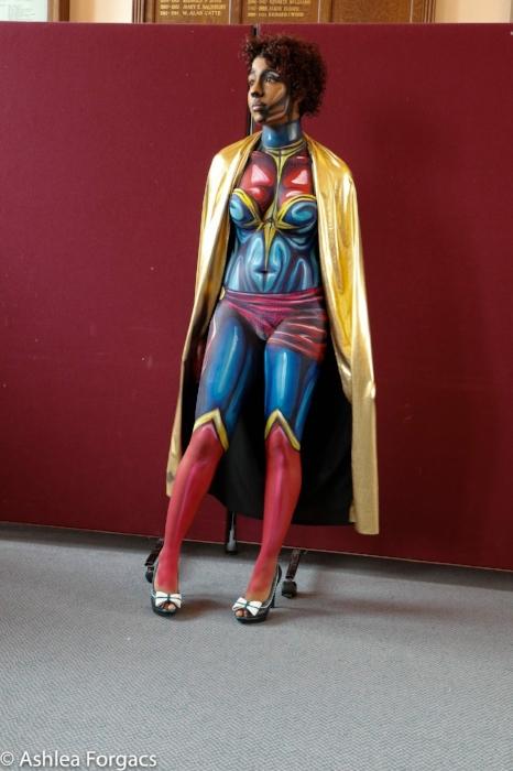 Rhyana posing with Ms. Marvel body paint in a gold cloak against a red background.