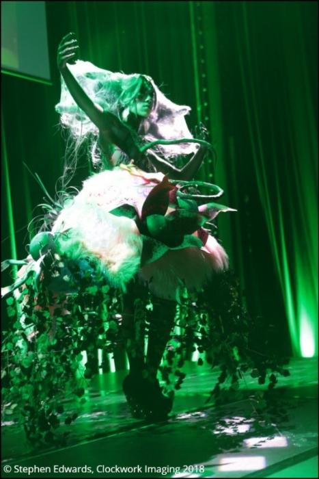 Rhyana in a forest-themed outfit with arms outstretched on a stage in green lighting.