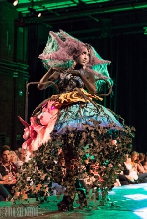 Rhyana in a forest-themed outfit with arms held together, walking on a modelling catwalk in green lighting.