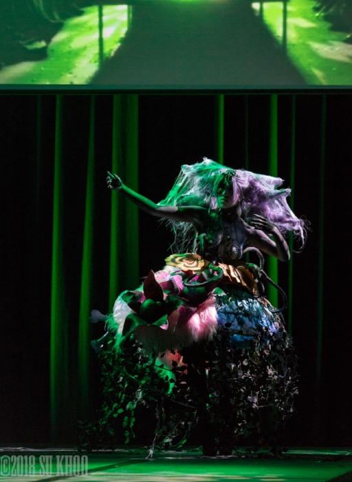 Rhyana in a forest-themed outfit against a black and green striped background.