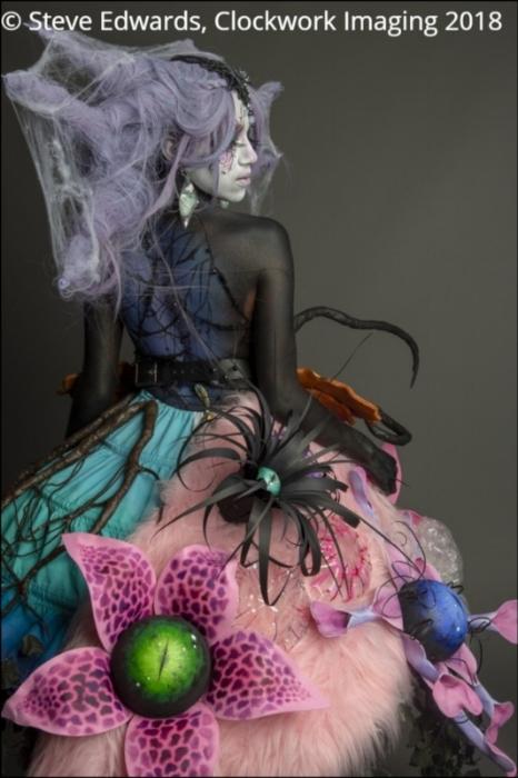 Rhyana in a spooky forest-themed outfit leaning backwards over their shoulder against a grey background.