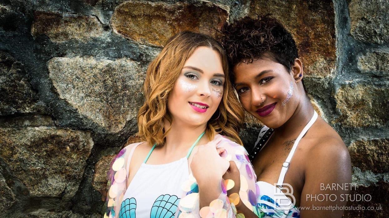 Rhyana and Megan smiling and leaning against each other in front of a brown and grey cobblestone wall.