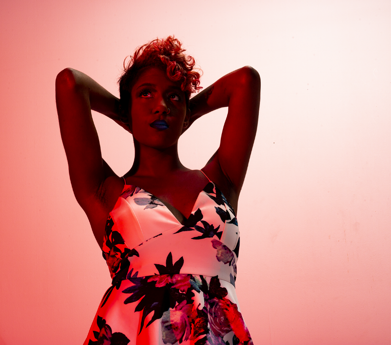 Rhyana in a white floral dress with both hands behind their head, looking upwards in red lighting.