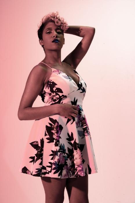 Rhyana looking towards the camera in a white floral dress with one hand behind their head.