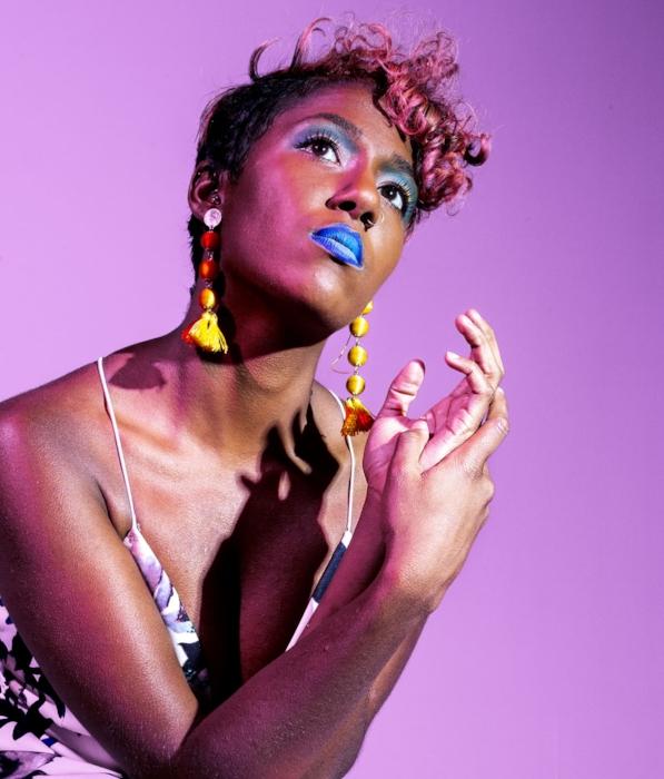 Rhyana against a purple background with hands clasped in front of them, looking upwards off camera.