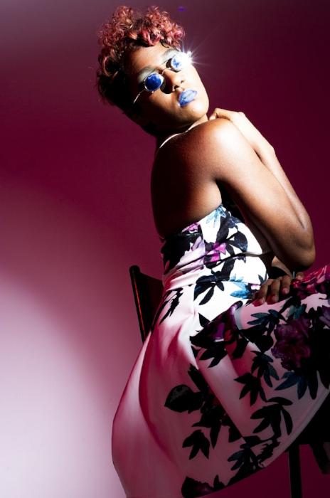 Rhyana in a white floral dress with blue lipstick and sunglasses, looking over their shoulder at the camera.