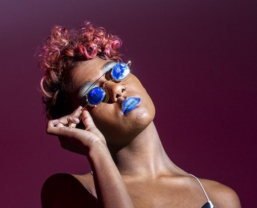 Rhyana in blue-lensed sunglasses and matching lipstick, one hand on the frames as they look off camera.