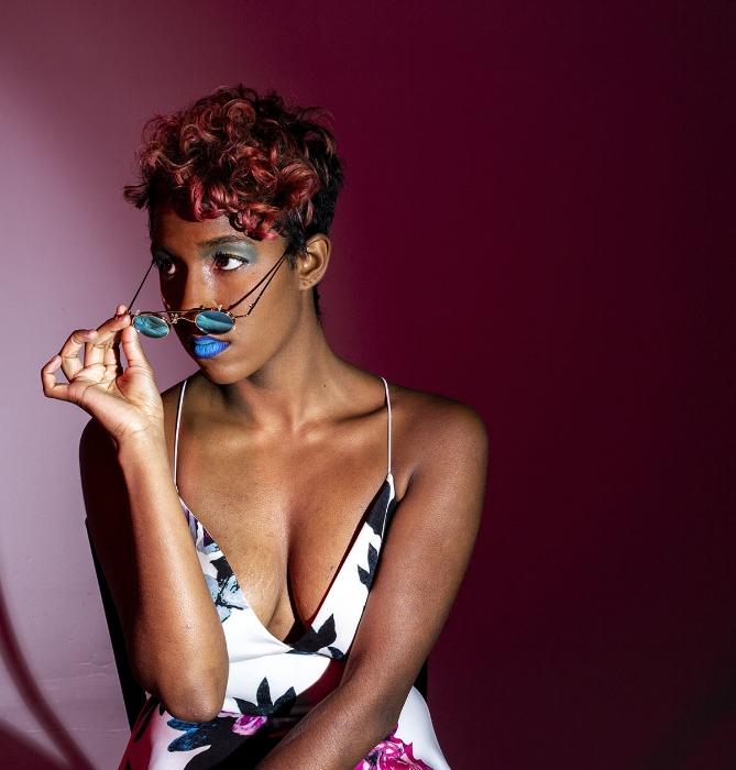 Rhyana sat in a white floral dress with blue lipstick, peering off camera over the rim of blue-lensed sunglasses.