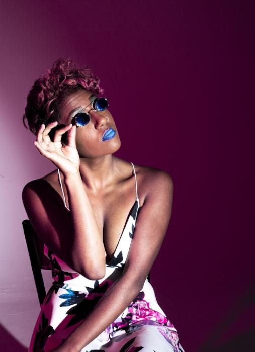 Rhyana sat in a whie floral dress with one hand on their sunglasses, looking off camera.