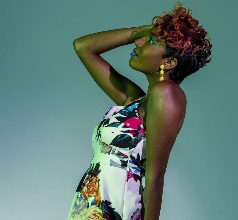 Rhyana leaning back with one hand on their face in a white floral dress against a green background.