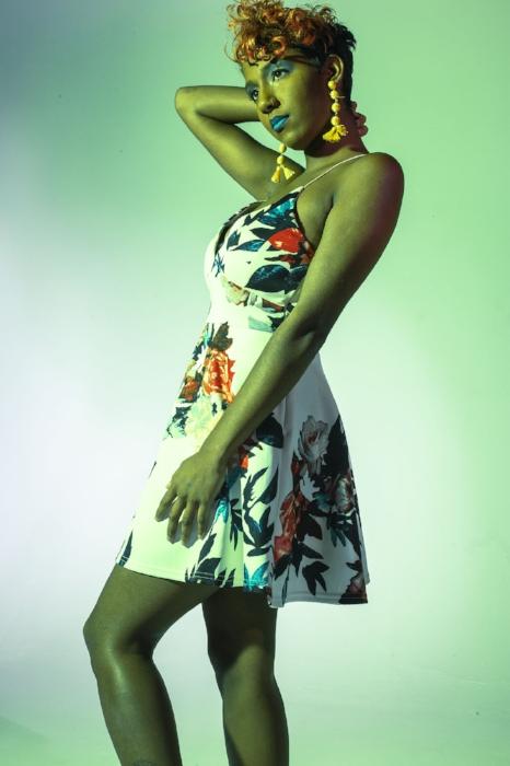 Rhyana with one hand on their face in a white floral dress and blue lipstick, stood in green lighting.