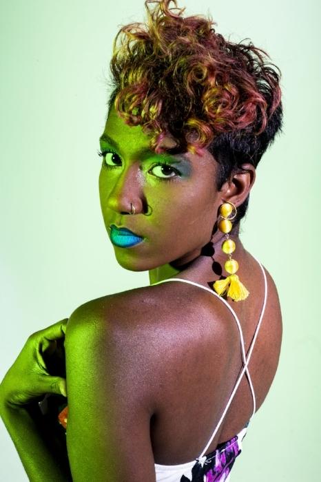 Rhyana looking back over their shoulder in blue lipstick with yellow tasseled-earing, looking towards camera.