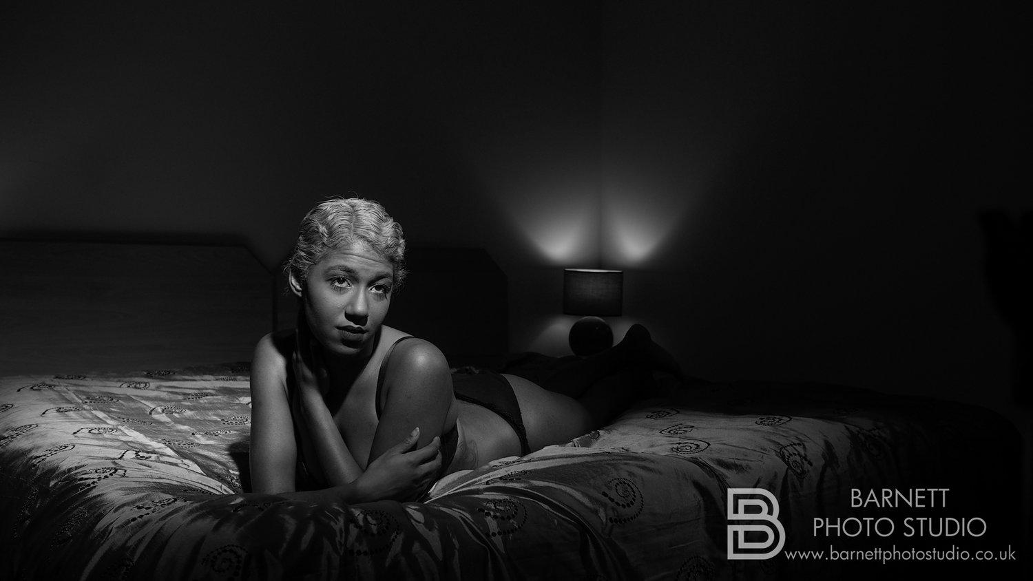 Black and white shot of Rhyana wearing lingerie, lying on their front in bed with their arms crossed.