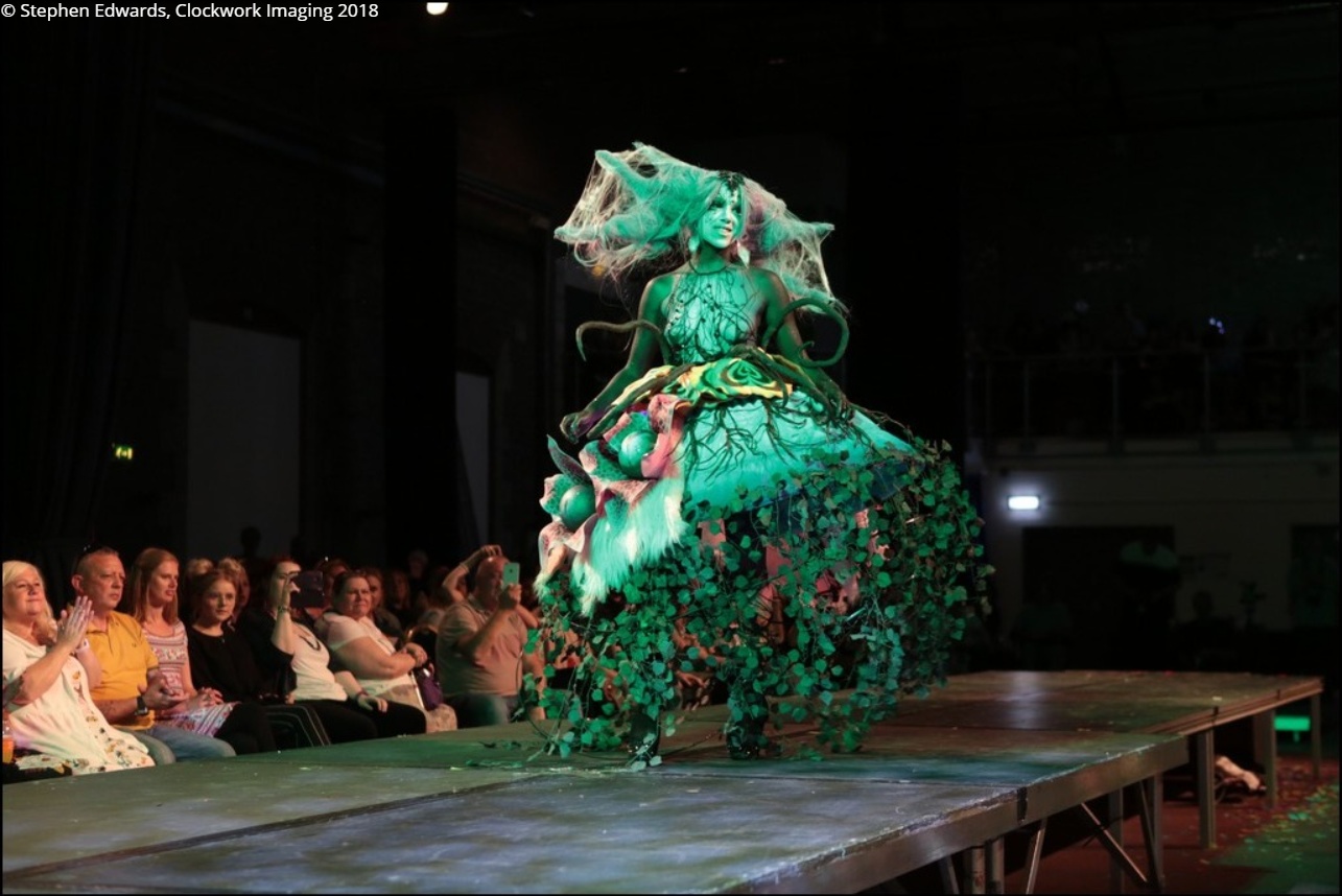 Long shot of Rhyana in a forest-themed outfit with green lighting on a modelling catwalk.