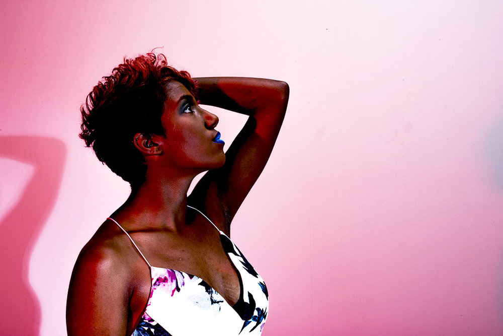 Rhyana in a white floral dress with one arm on her head, looking off camera against a pink background.